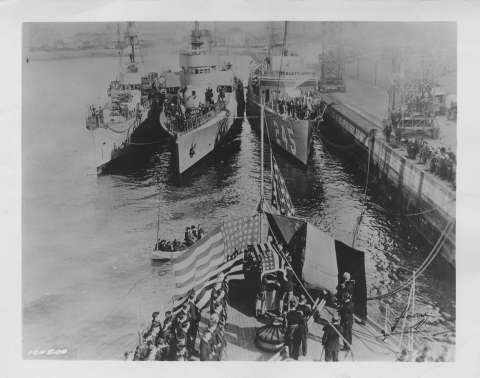 Installation du cercueil sur le catafalque à la poupe de l’US cruiser Olympia. En arrière-plan l’escorte navale française et américaine : le contre-torpilleur Amiral-Sénès, le torpilleur M.P.-Cestin et l’US destroyer Reuben-James